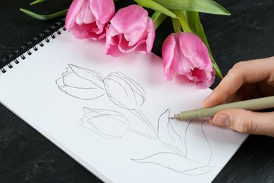 Photo of Woman sketching tulips in notebook at black table, closeup