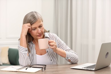 Photo of Menopause. Tired woman with cup of drink at wooden table indoors
