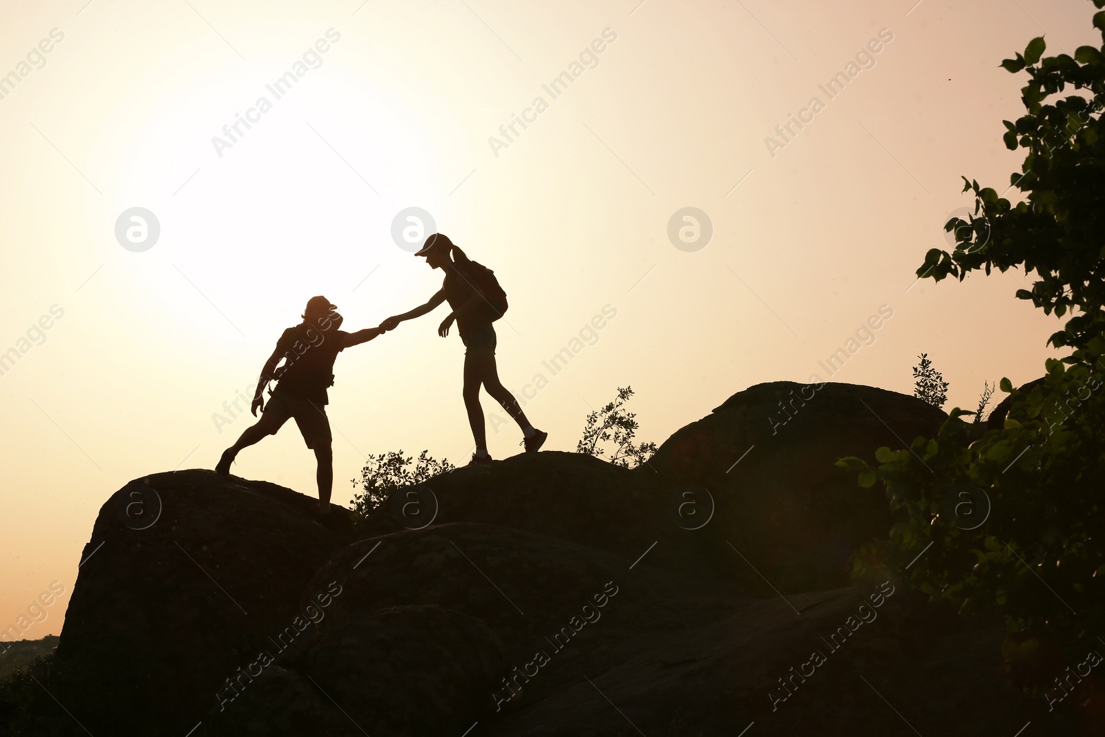 Photo of Silhouettes of man and woman helping each other to climb on hill against sunset