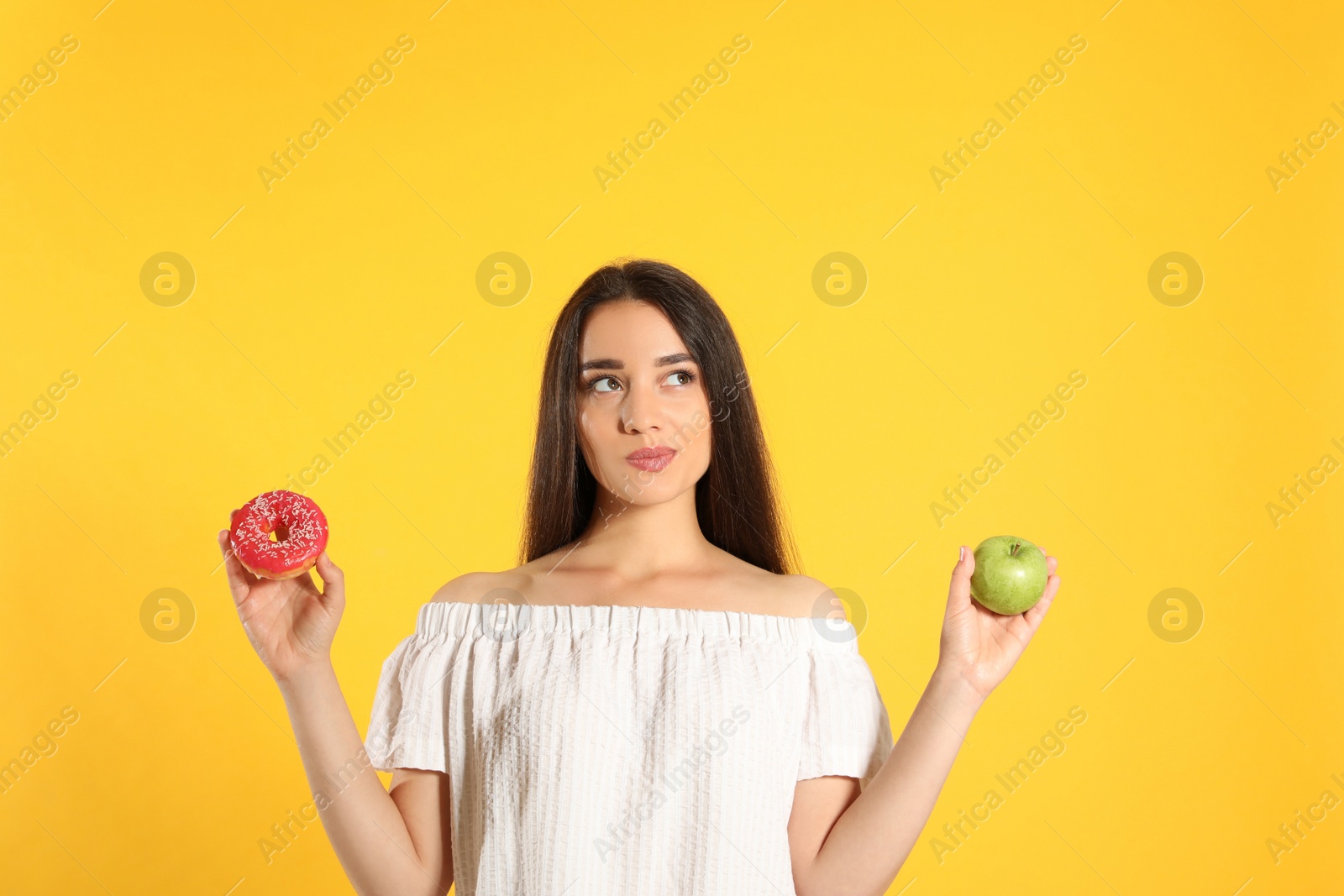 Photo of Doubtful woman choosing between apple and doughnut on yellow background