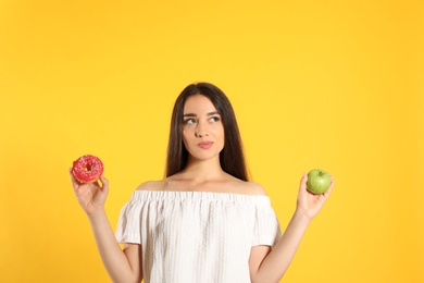 Doubtful woman choosing between apple and doughnut on yellow background