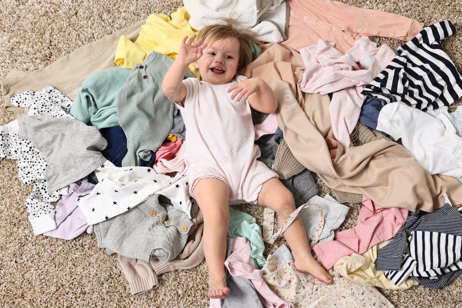 Photo of Little girl and pile of baby clothes on carpet, above view