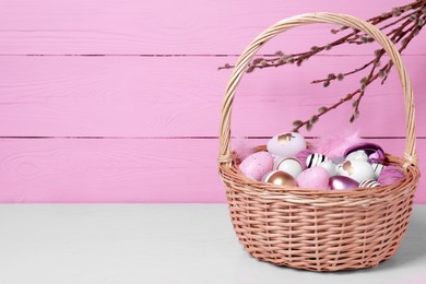 Photo of Wicker basket with festively decorated Easter eggs on white table against pink wooden background. Space for text