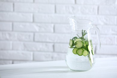 Photo of Refreshing cucumber water with rosemary in jug on white table against brick wall. Space for text