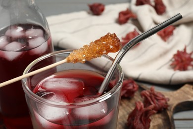 Refreshing hibiscus tea with ice cubes and stick with sugar crystals on table, closeup