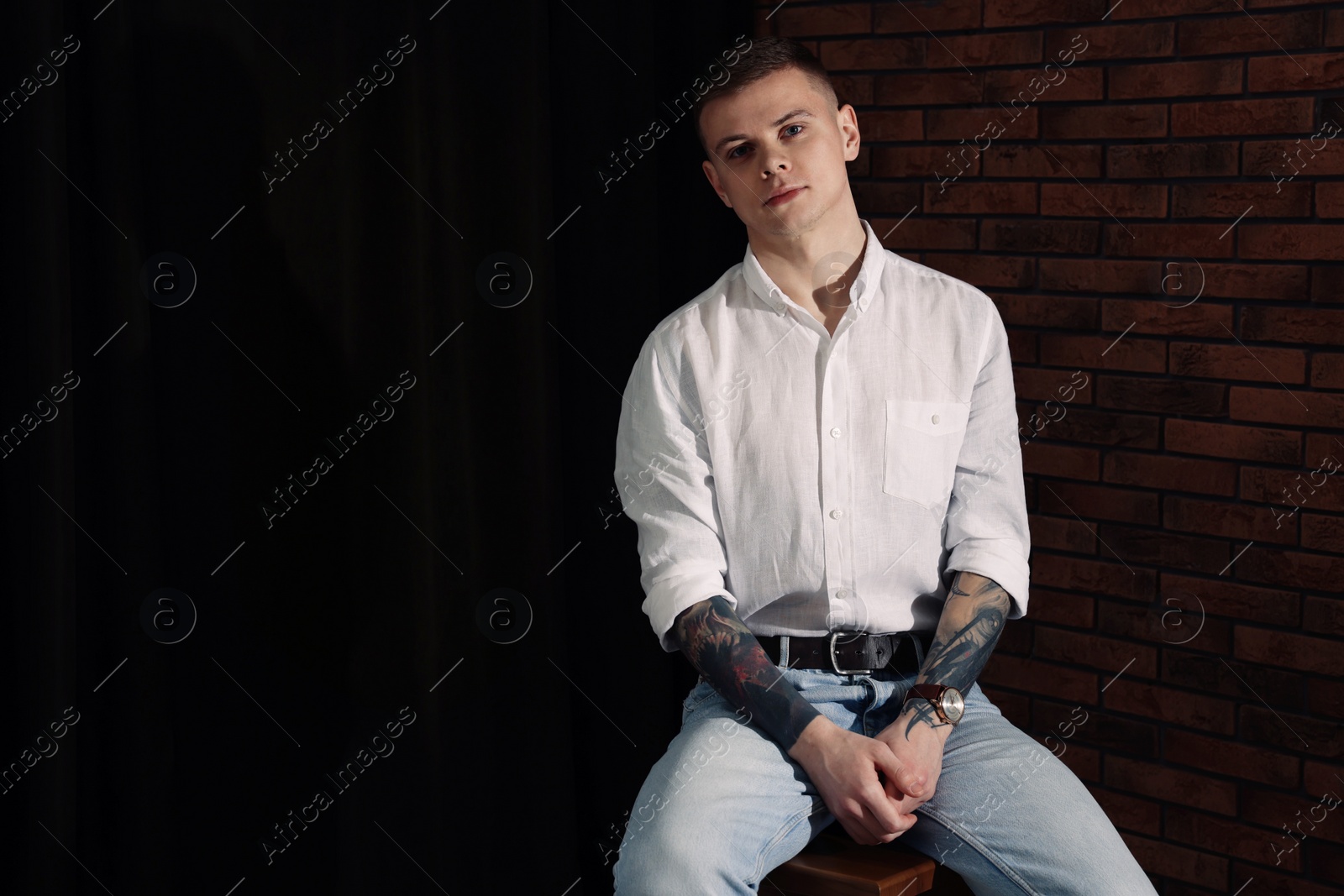 Photo of Young man with tattoos sitting on stool indoors. Space for text