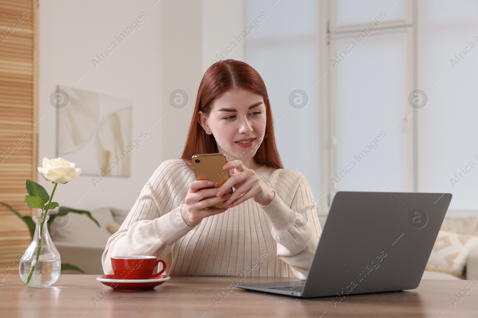 Photo of Beautiful woman with smartphone using laptop at wooden table in room