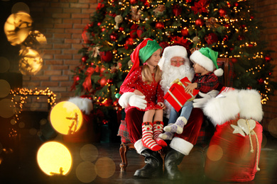 Photo of Santa Claus and little children with present near Christmas tree indoors
