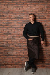 Photo of Full length portrait of happy young waiter in uniform near brick wall indoors