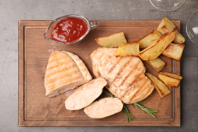 Photo of Wooden board with grilled meat, garnish and sauce on grey background, top view