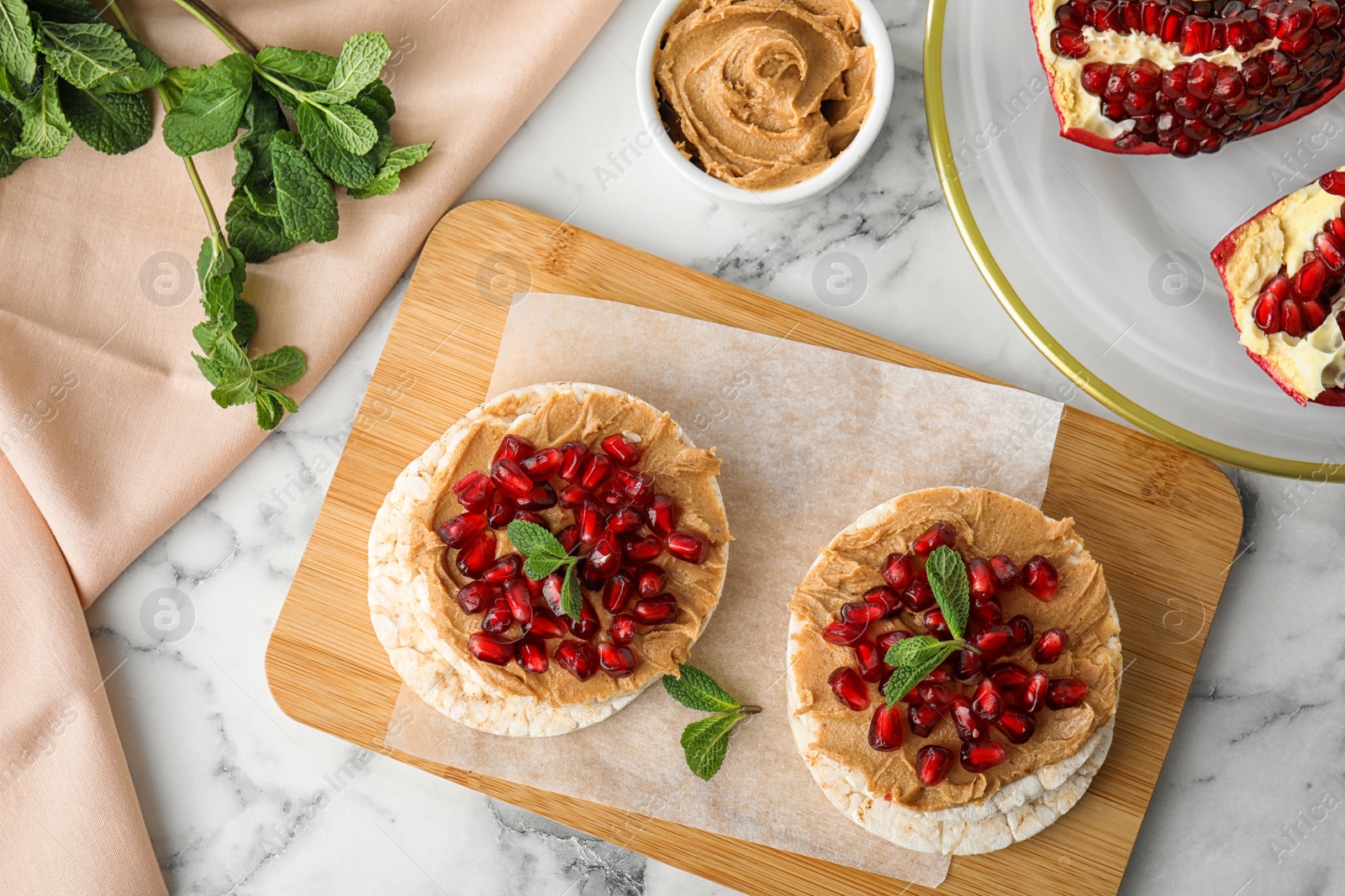 Photo of Puffed rice cakes with peanut butter, pomegranate seeds and mint on white marble table, flat lay