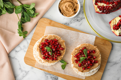 Photo of Puffed rice cakes with peanut butter, pomegranate seeds and mint on white marble table, flat lay