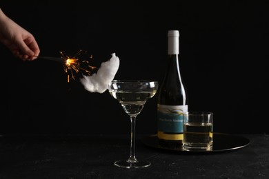 Photo of Cocktail with tasty cotton candy and bottle of alcohol drink on dark textured table. Woman holding sparkler against black background, closeup