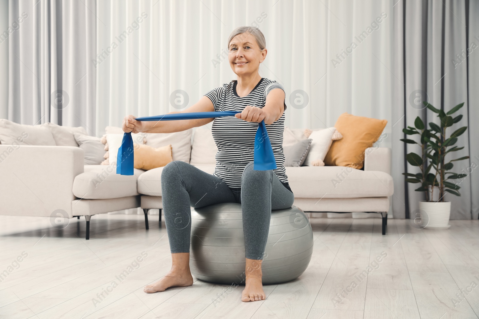 Photo of Senior woman doing exercise with elastic resistance band on fitness ball at home