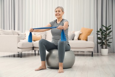 Photo of Senior woman doing exercise with elastic resistance band on fitness ball at home