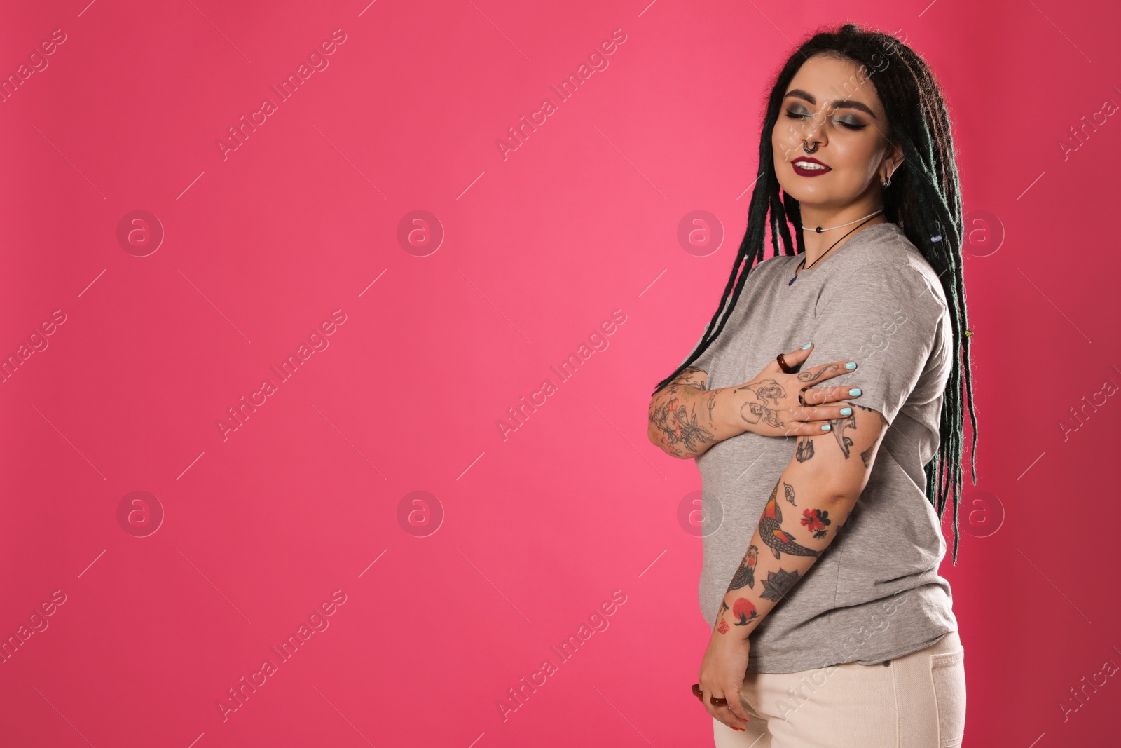 Photo of Beautiful young woman with tattoos on arms, nose piercing and dreadlocks against pink background. Space for text