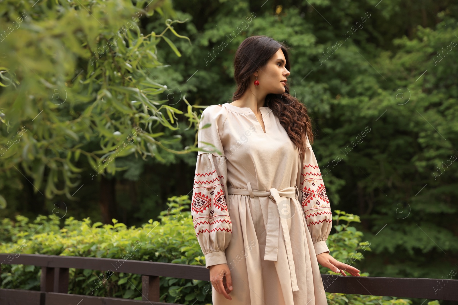 Photo of Beautiful woman wearing embroidered dress near wooden railing in countryside. Ukrainian national clothes