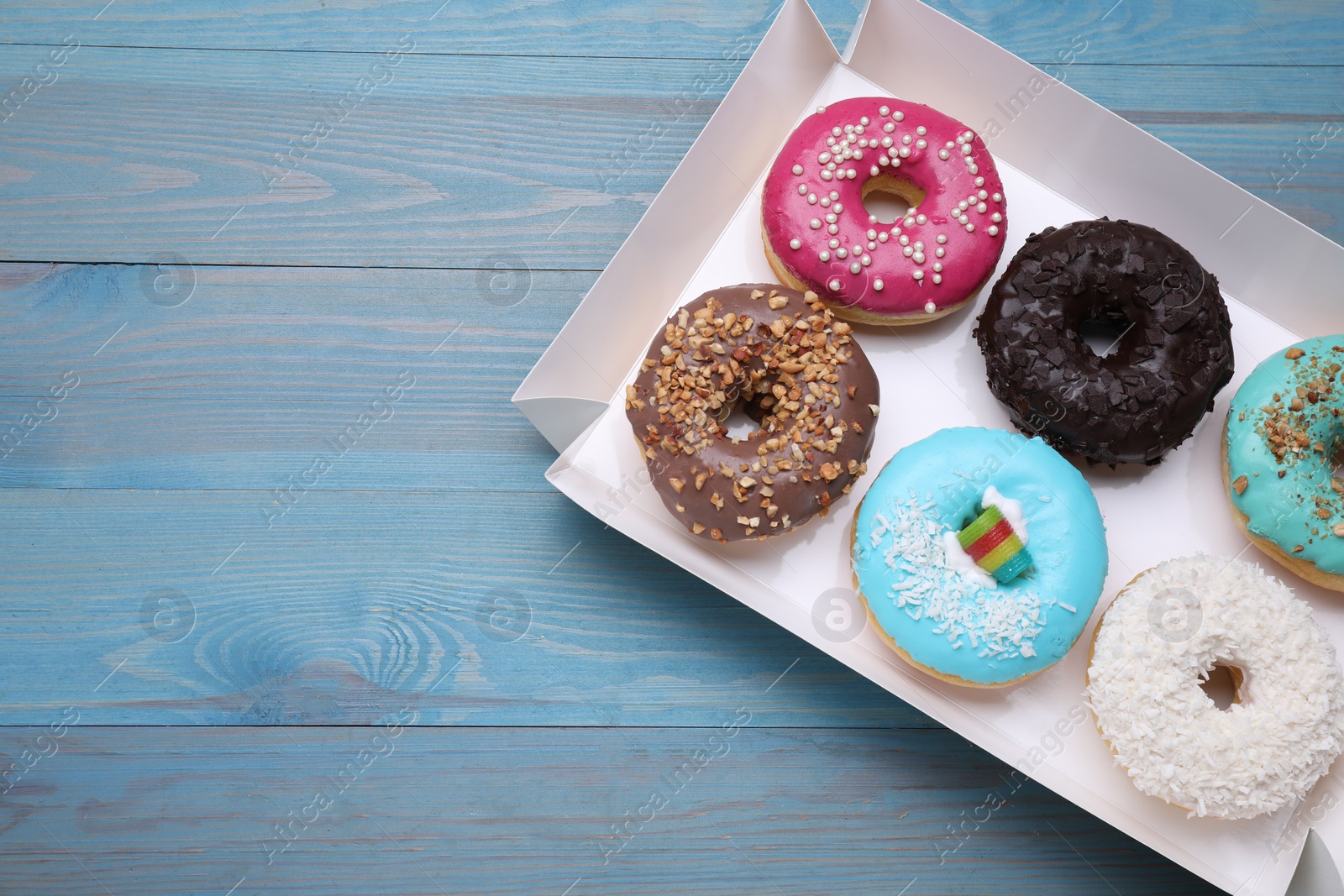 Photo of Box with different tasty glazed donuts on light blue wooden table, top view. Space for text