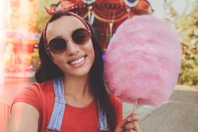 Stylish young woman with cotton candy taking selfie at funfair on sunny day