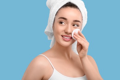 Photo of Beautiful woman in terry towel removing makeup with cotton pad on light blue background