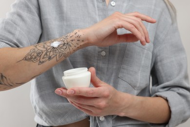 Photo of Tattooed woman with jar of cream on light background, closeup