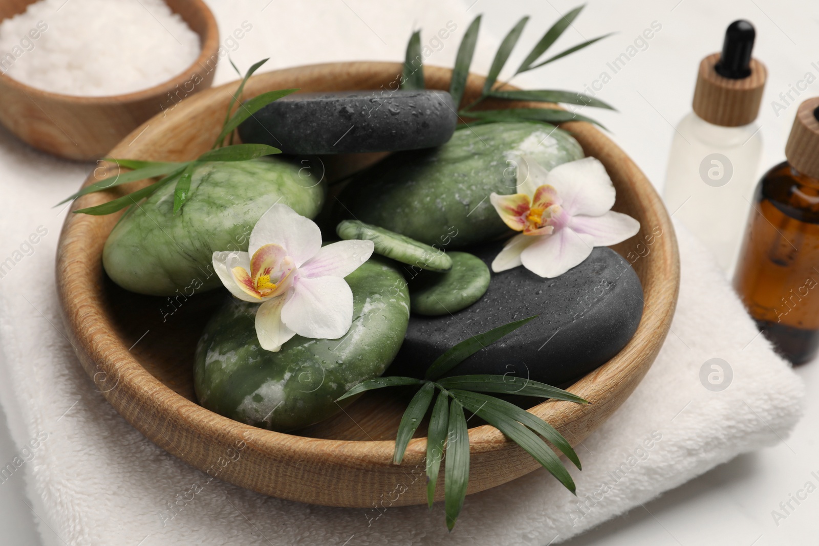 Photo of Spa stones, beautiful flowers and green leaves in wooden bowl on white marble table, closeup