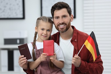 Photo of Immigration. Happy man with his daughter holding passports and flag of Germany indoors