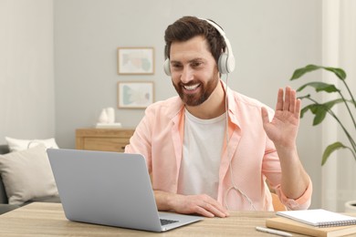 Photo of Man in headphones greeting someone during video chat via laptop at home