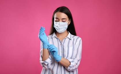 Woman in protective face mask putting on medical gloves against pink background