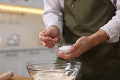 Photo of Making bread. Man putting salt into bowl with flour at table in kitchen, closeup