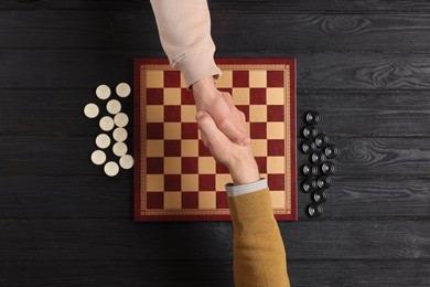 Photo of Playing checkers. Partners shaking hands after match at black wooden table, top view