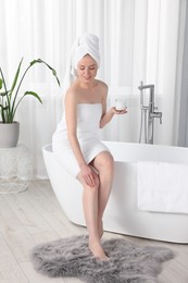 Photo of Beautiful young woman applying body cream onto leg in bathroom