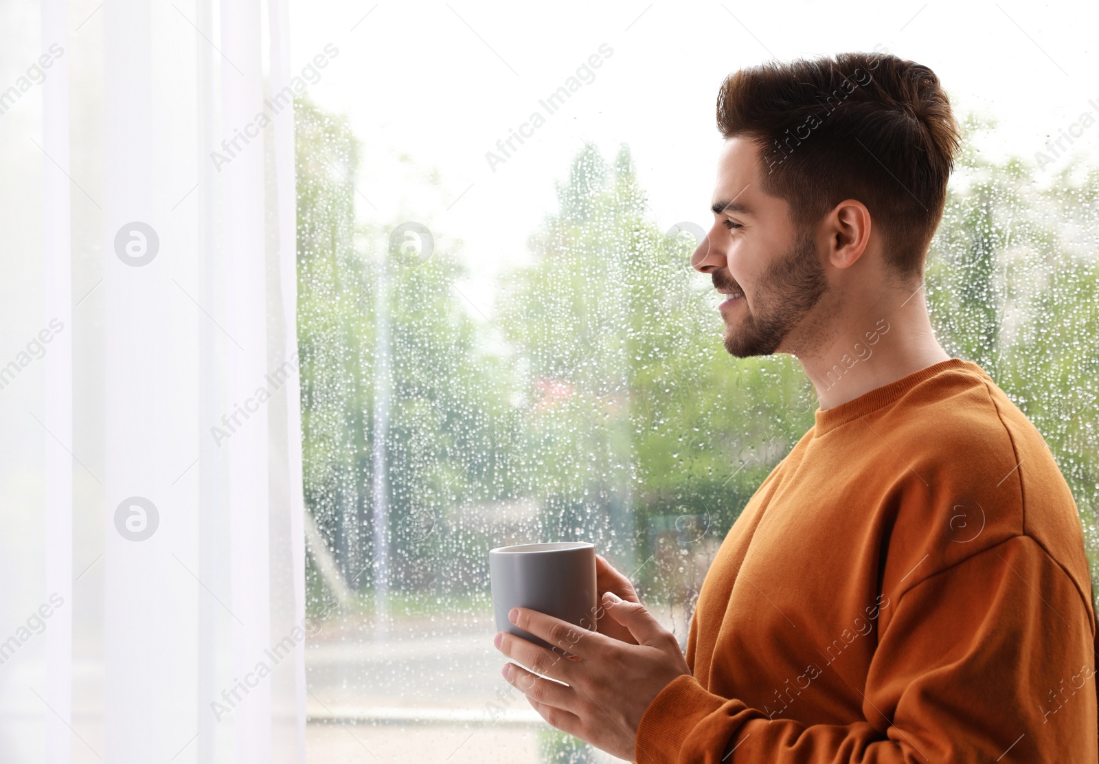 Photo of Happy handsome man with cup of coffee near window on rainy day