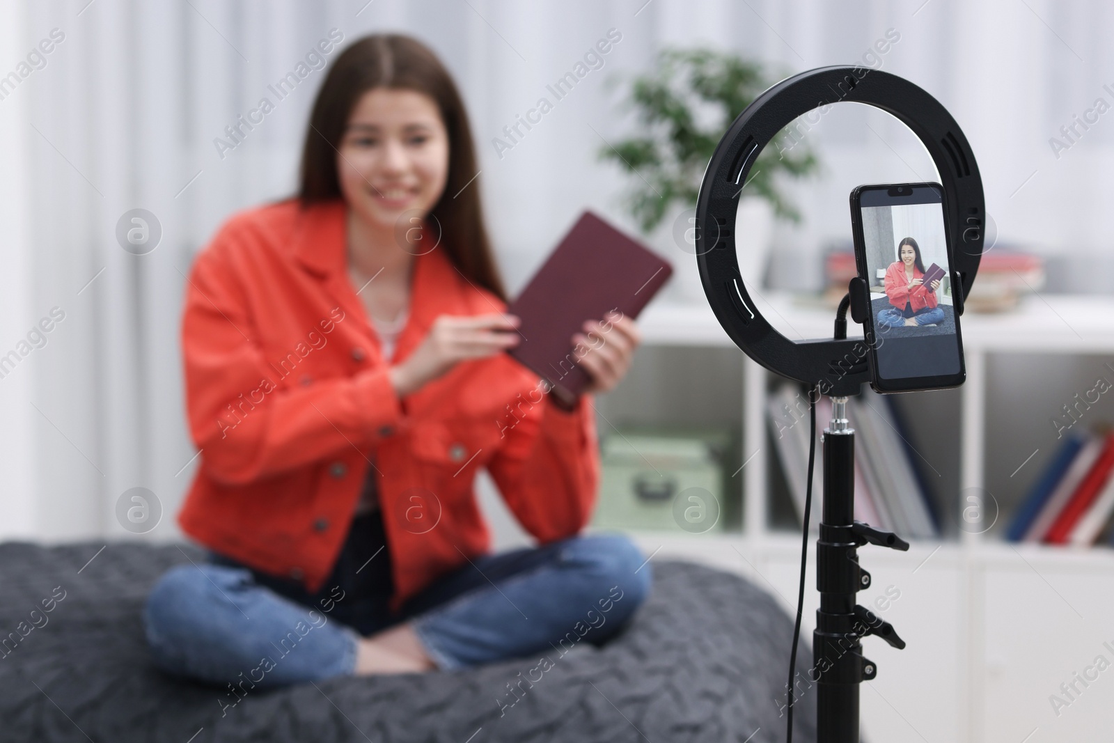 Photo of Teenage blogger with book streaming at home, focus on smartphone