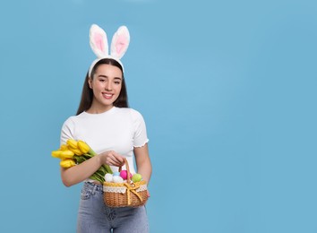 Photo of Happy woman in bunny ears headband holding wicker basket with painted Easter eggs and bouquet of flowers on turquoise background. Space for text