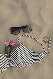 Beautiful sunglasses, swimsuit and tropical flower on sand, above view