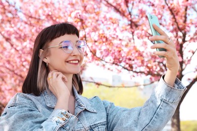 Photo of Beautiful young woman taking selfie in park with blossoming sakura trees