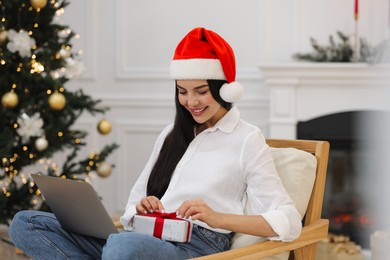 Celebrating Christmas online with exchanged by mail presents. Smiling woman in Santa hat opening gift box during video call on laptop at home