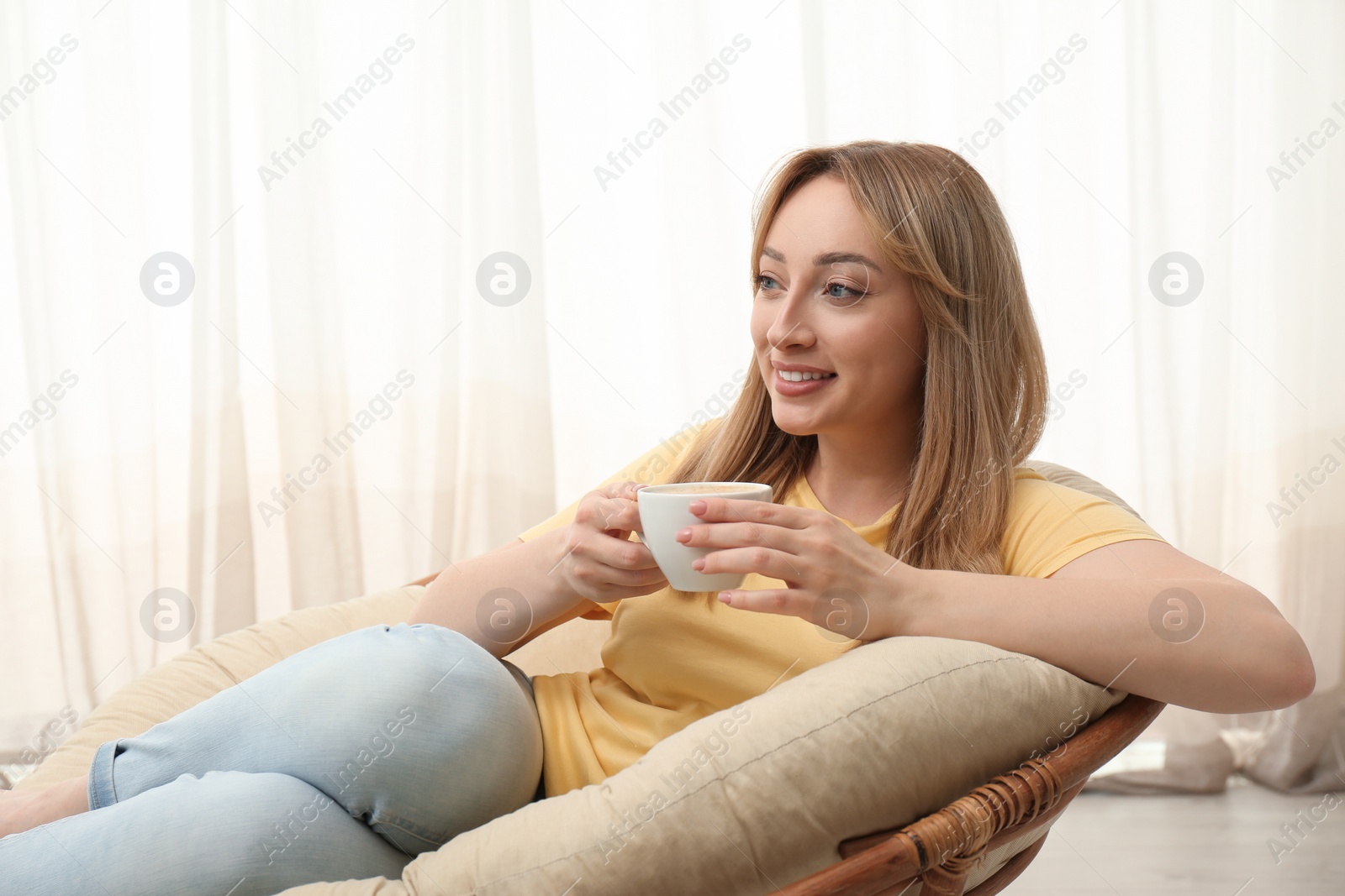 Photo of Beautiful young woman with cup of hot drink relaxing in papasan chair indoors