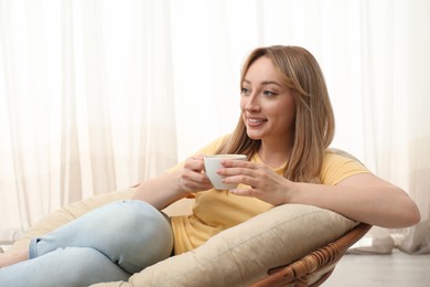 Beautiful young woman with cup of hot drink relaxing in papasan chair indoors