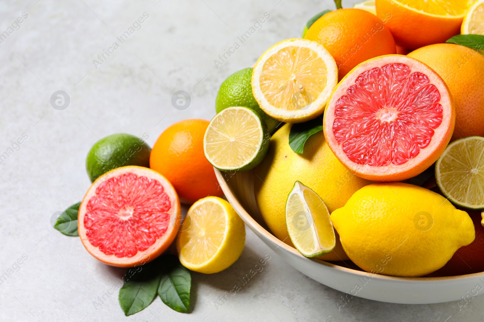 Photo of Different fresh citrus fruits and leaves in bowl on light table, closeup