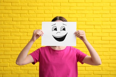 Woman hiding behind sheet of paper with laughing face against yellow brick wall