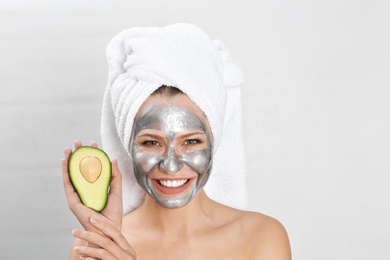 Beautiful woman holding avocado near her face with silver mask against light background. Space for text