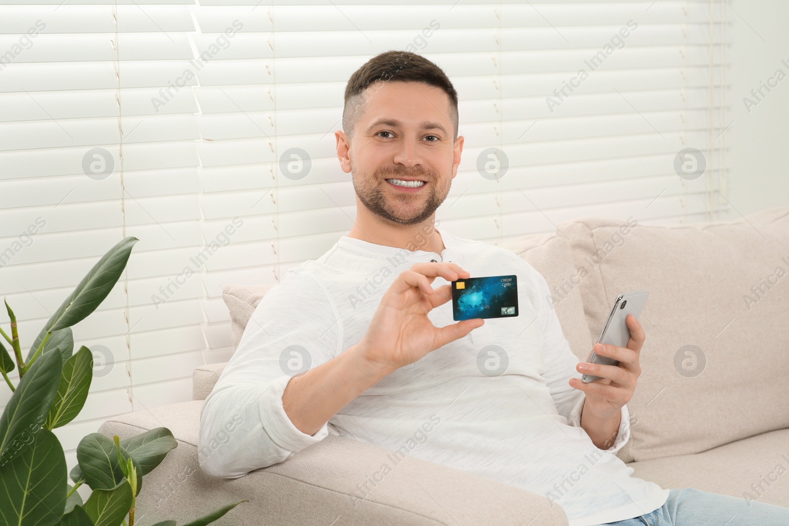 Photo of Happy man with credit card using smartphone for online shopping on sofa at home