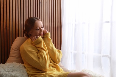 Beautiful young woman in warm sweater near window at home, space for text. Winter season