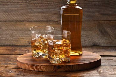 Photo of Whiskey with ice cubes in glasses and bottle on wooden table, closeup