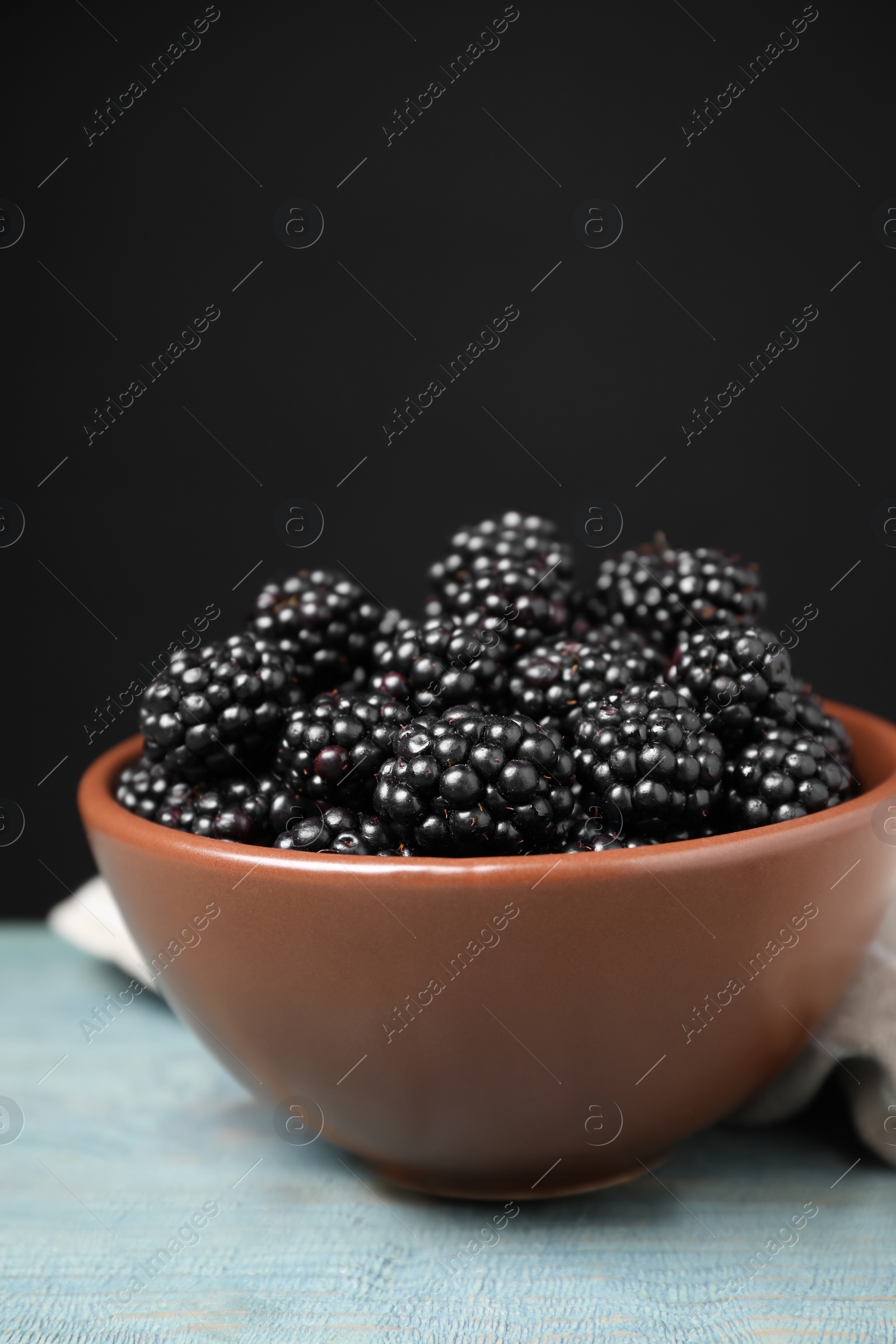 Photo of Fresh ripe blackberries in bowl on blue wooden table against dark background, closeup. Space for text