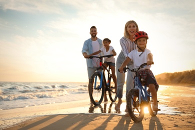 Photo of Happy parents teaching children to ride bicycles on sandy beach near sea at sunset
