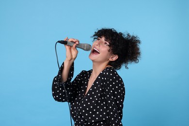 Beautiful young woman with microphone singing on light blue background