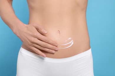 Photo of Woman applying body cream onto her belly against light blue background, closeup